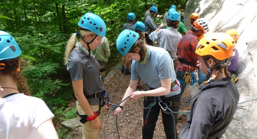 A group of students wearing helmets stand at the bottom of a rock wall. In the foreground, one person helps another tie their climbing ropes. 